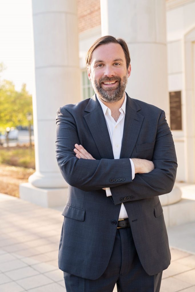 Justin Dion, Dallas LGBTQ+ therapist, standing with arms crossed and smiling confidently outdoors in a navy suit, with white columns and warm sunlight in the background.