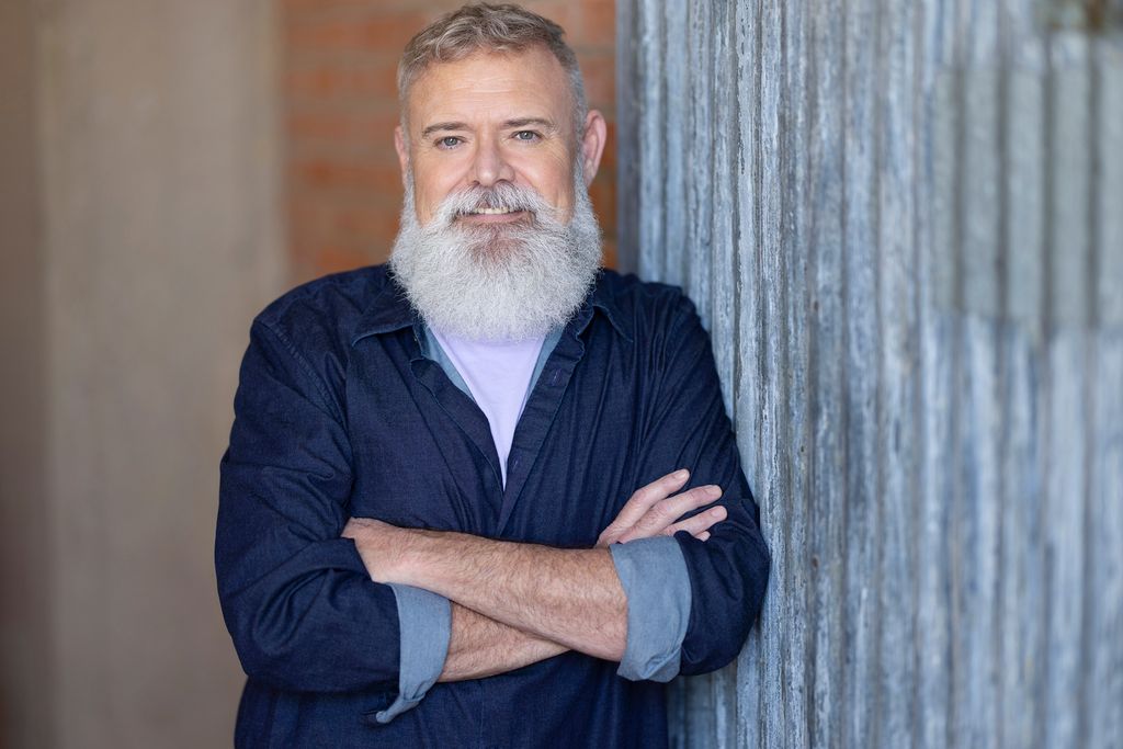 A male therapist with short gray hair and a full white beard, wearing a navy blue button-down shirt, standing with arms crossed and leaning against a weathered pillar, smiling warmly at the camera.