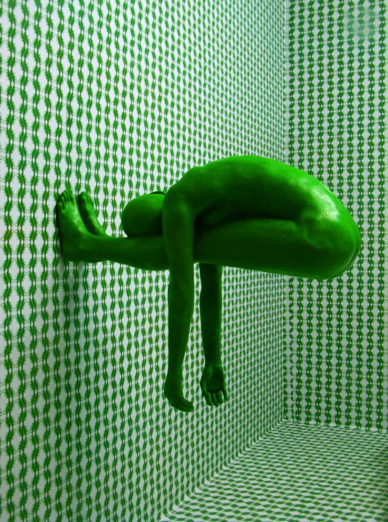 Green-tinted human figure curled tightly into a corner of a patterned green room, body pressed against the wall in a protective posture, visually representing chronic stress, vigilance, and bodily containment.
