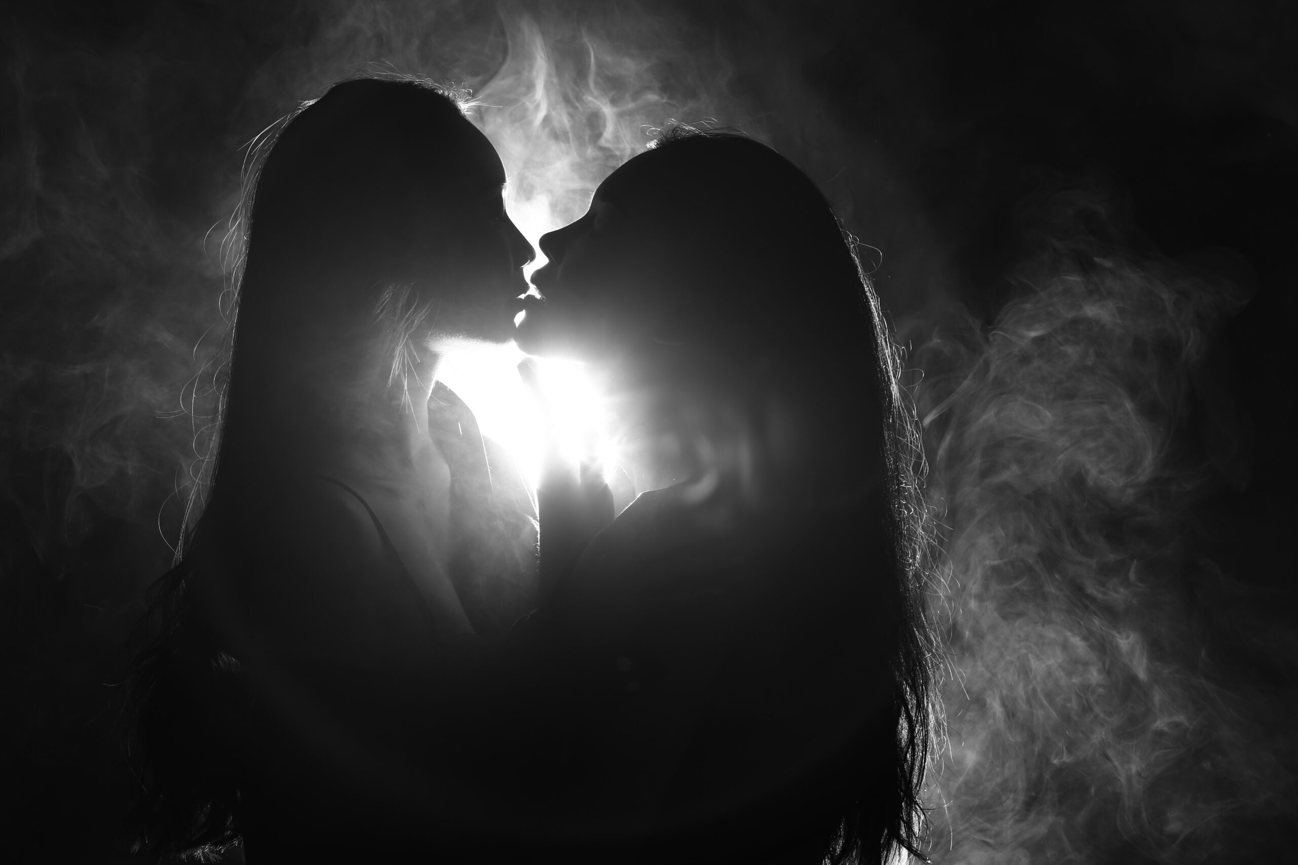 Black and white silhouette of two people leaning close together, faces nearly touching, with a bright light glowing between them and soft wisps of smoke surrounding them.