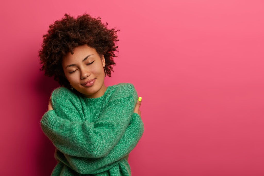 Person with eyes closed hugging themselves, wearing a green sweater against a pink background.
