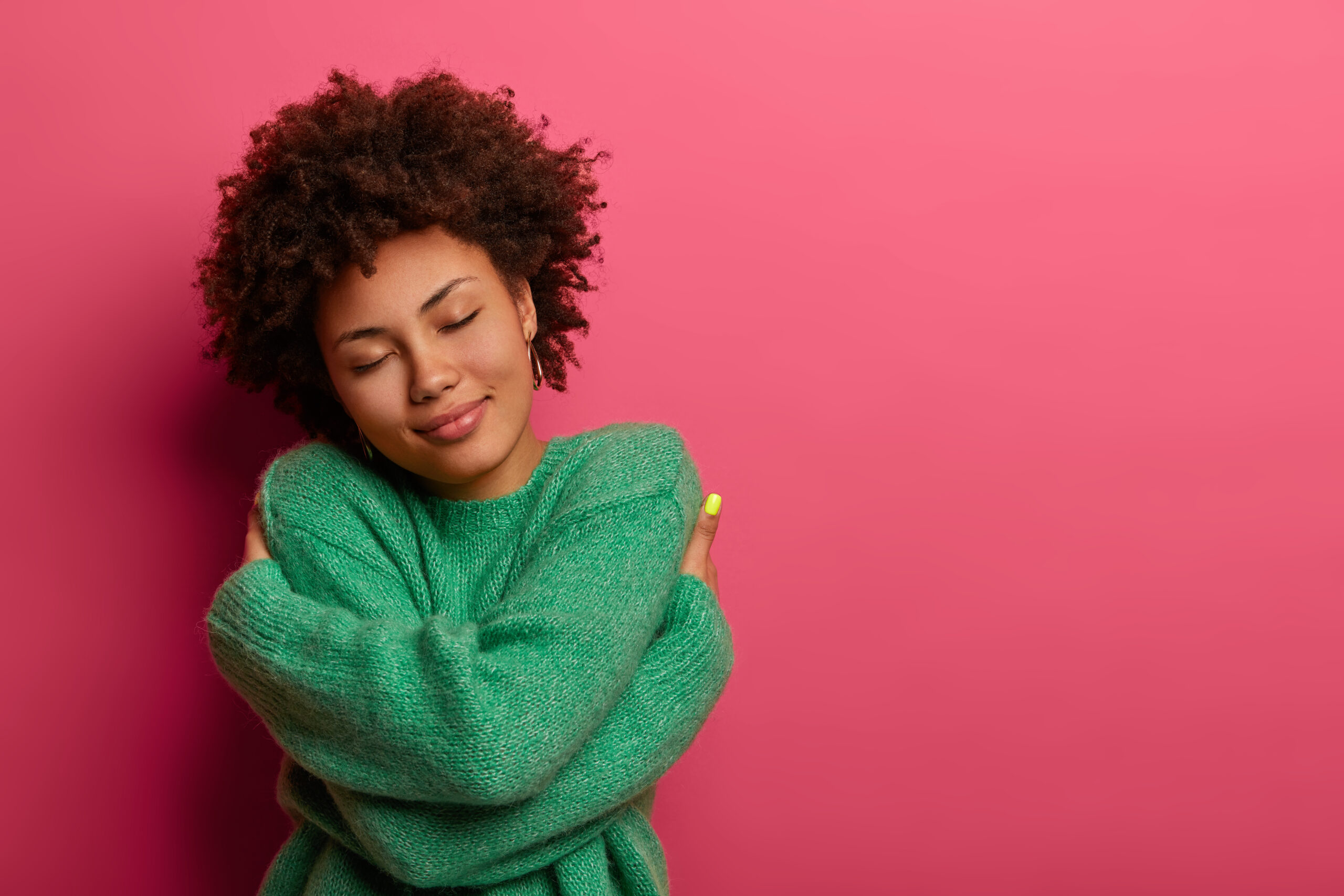 Person with eyes closed hugging themselves, wearing a green sweater against a pink background.