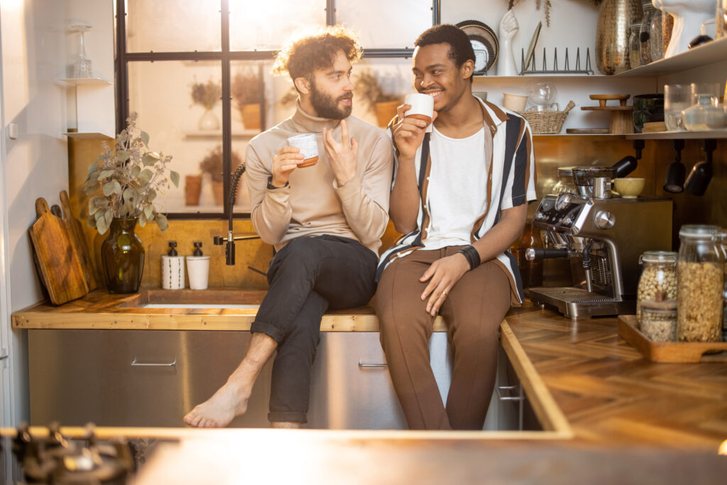 Two men sit together on a kitchen counter, holding mugs and talking comfortably in a warm, softly lit home kitchen.