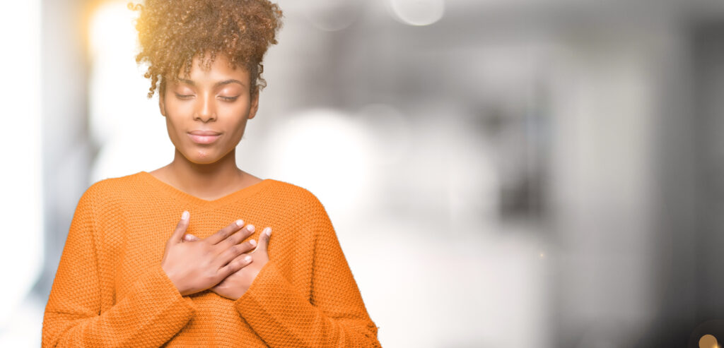 Black woman in orange sweater with eyes closed and hands resting over heart in calm self-compassion pose