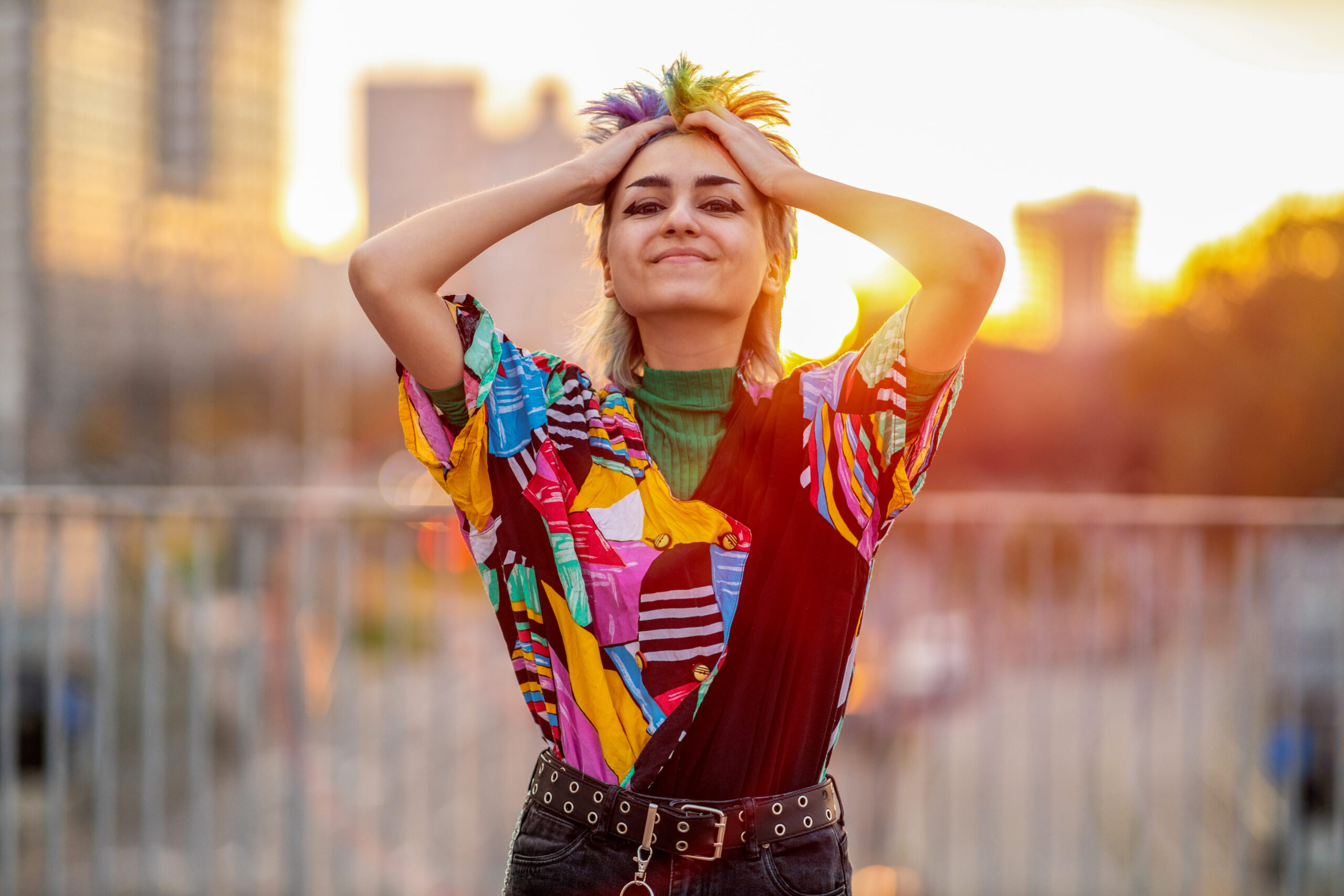A young person with multicolored hair smiles with hands resting on their head, standing outdoors in golden hour light wearing a vibrant patchwork shirt, conveying ease and self-possession.