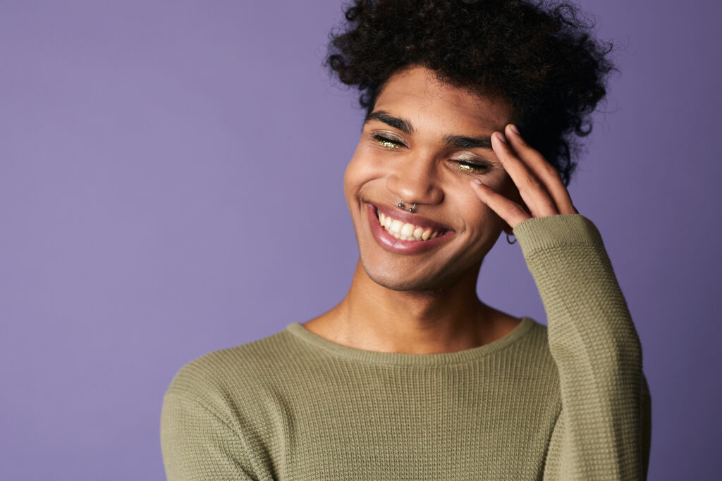 A young person of color with curly hair, glitter eye makeup, and a nose ring laughs joyfully against a purple background, representing gender euphoria and trans joy in LGBTQ+ affirming mental health care.