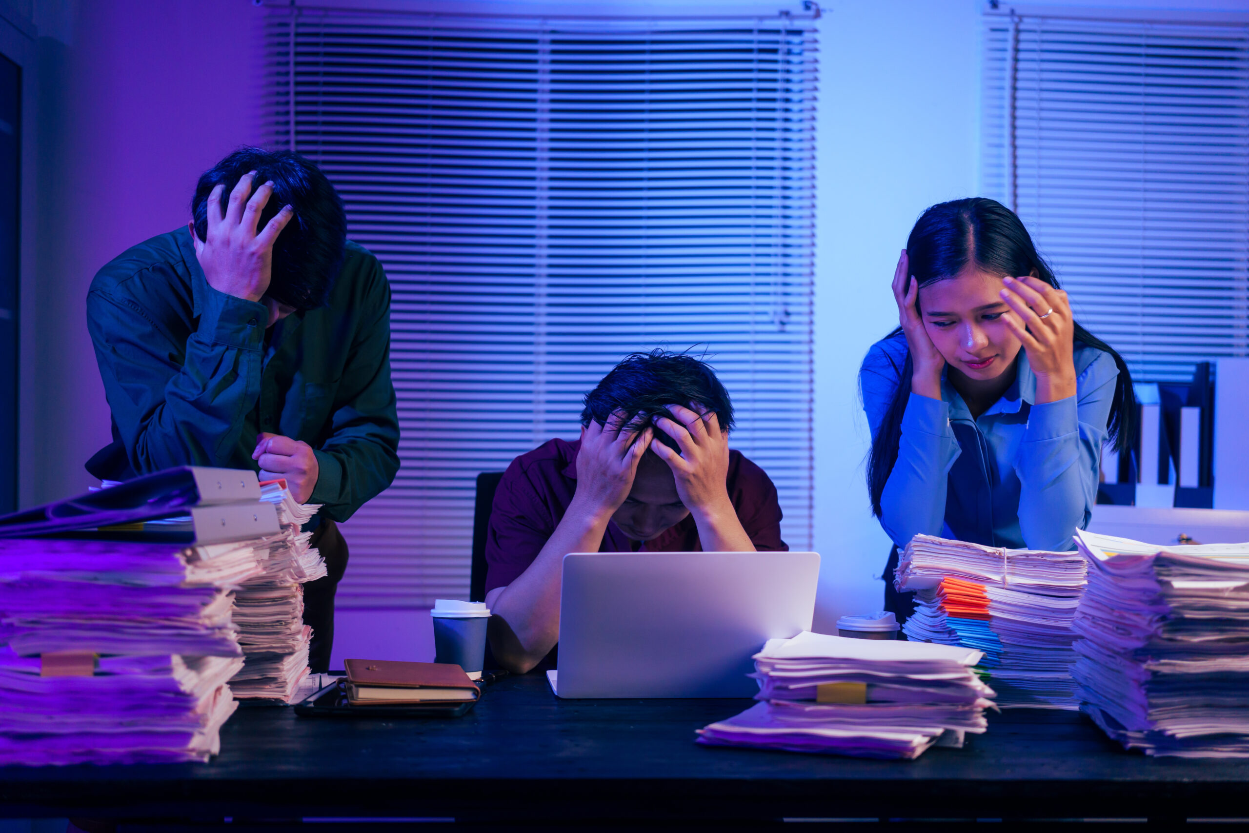 Three overwhelmed office workers surrounded by stacks of paperwork in a dimly lit, blue-toned office, heads in hands — illustrating workplace stress and nervous system dysregulation