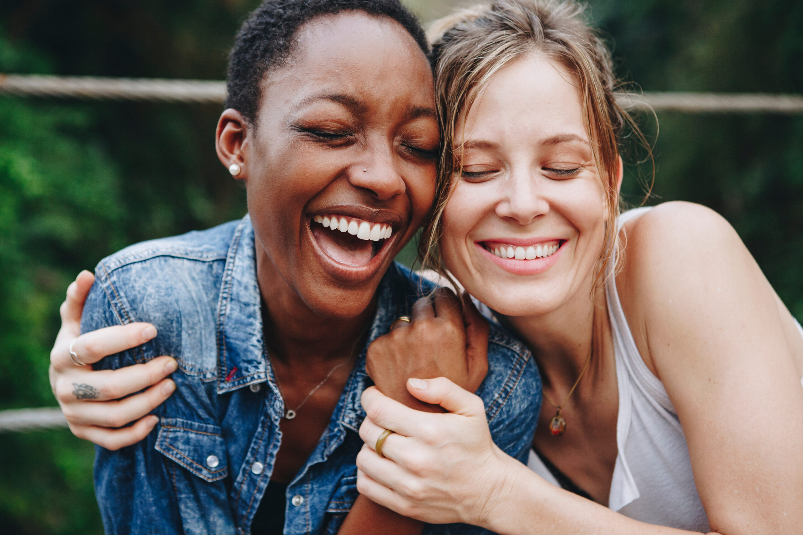 Two women, one Black and one white, embrace and laugh together outdoors with eyes closed, green trees visible in the background.