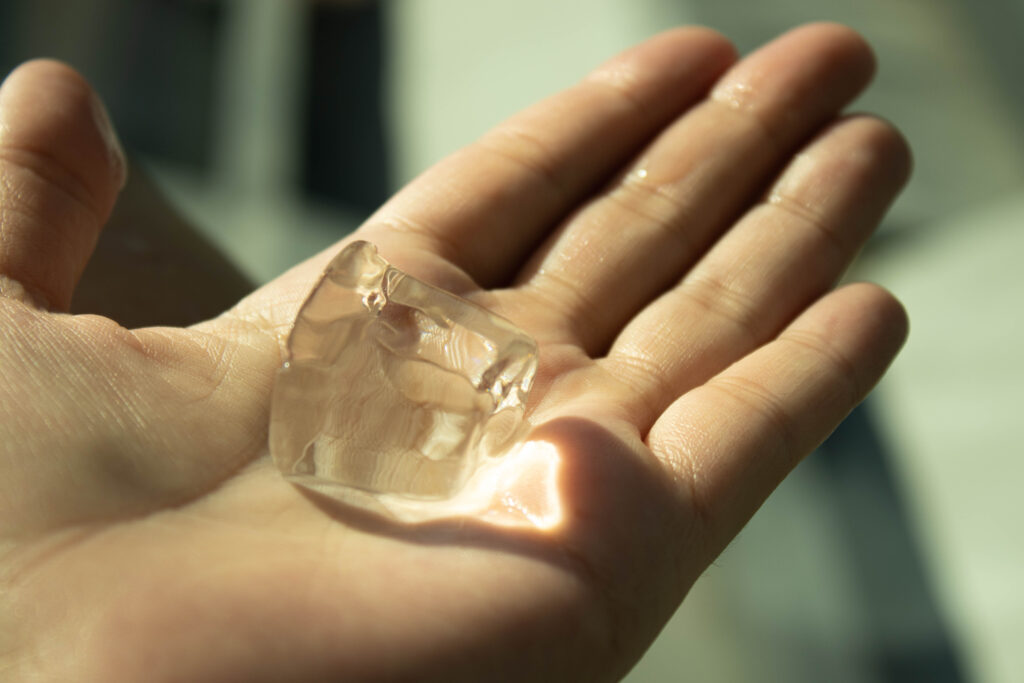 A close-up of an open palm holding a melting ice cube. Water droplets glisten across the skin as the ice catches warm light from below, creating a translucent amber glow against the hand.