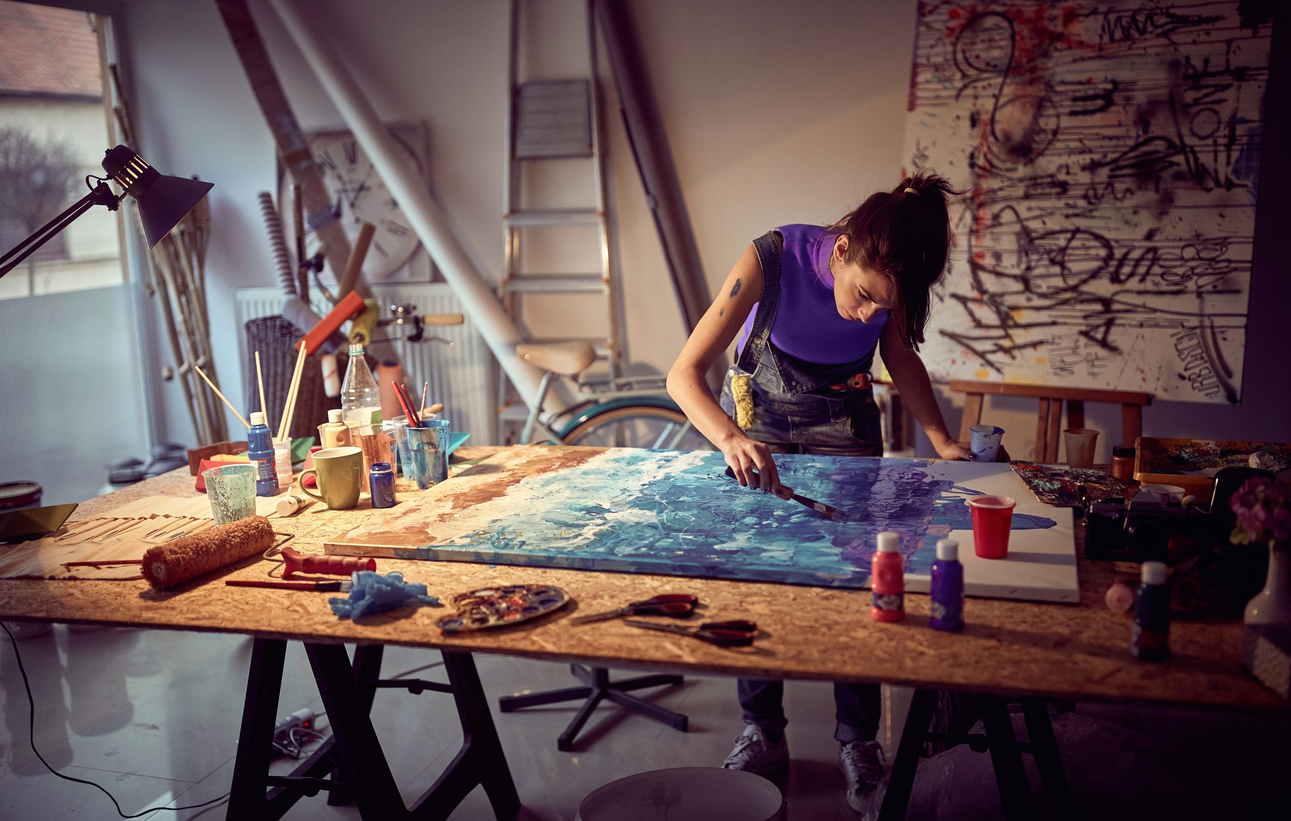 Artist painting a large abstract blue canvas in a softly lit studio filled with brushes, paint bottles, sketches, and creative tools