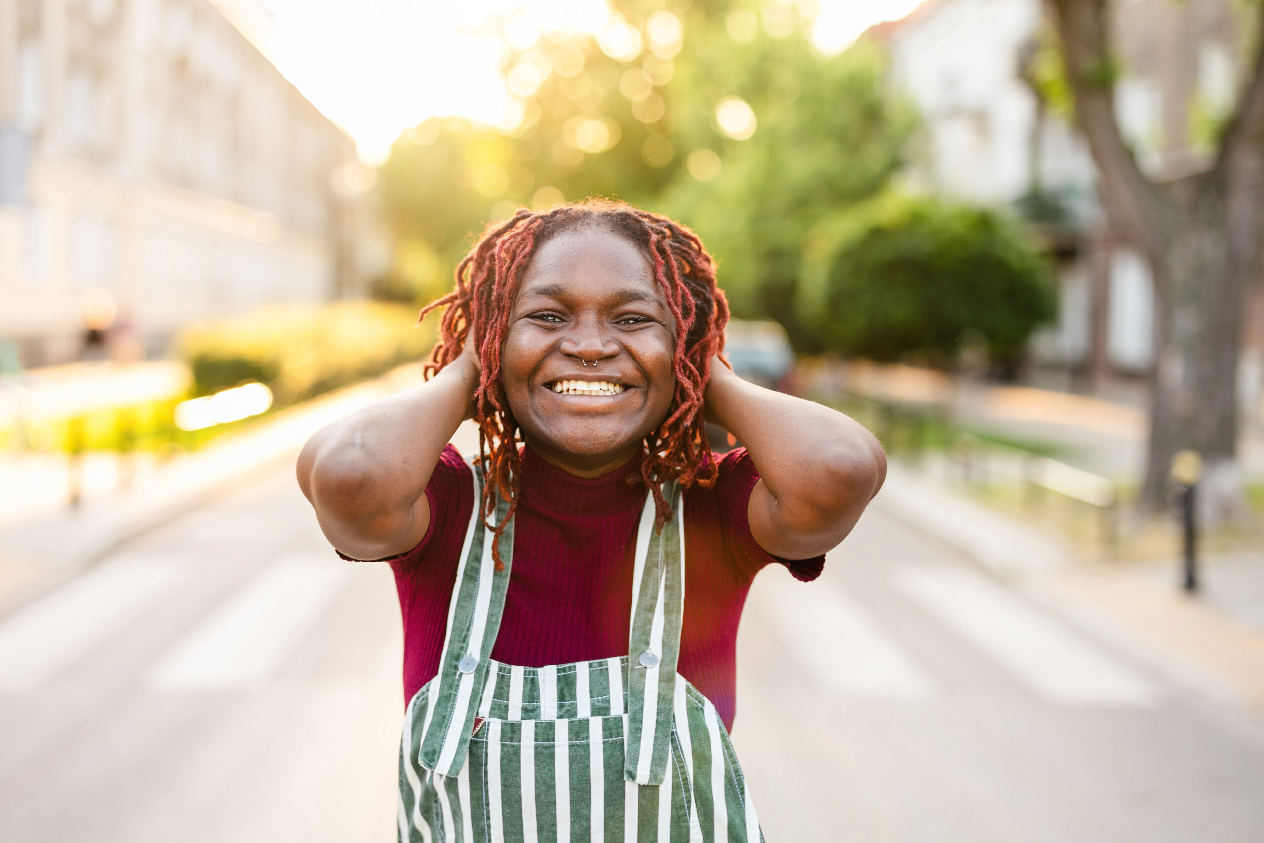 A Black woman with red locs and a nose ring laughs freely outdoors in striped overalls, hands behind her head, golden sunlight behind her