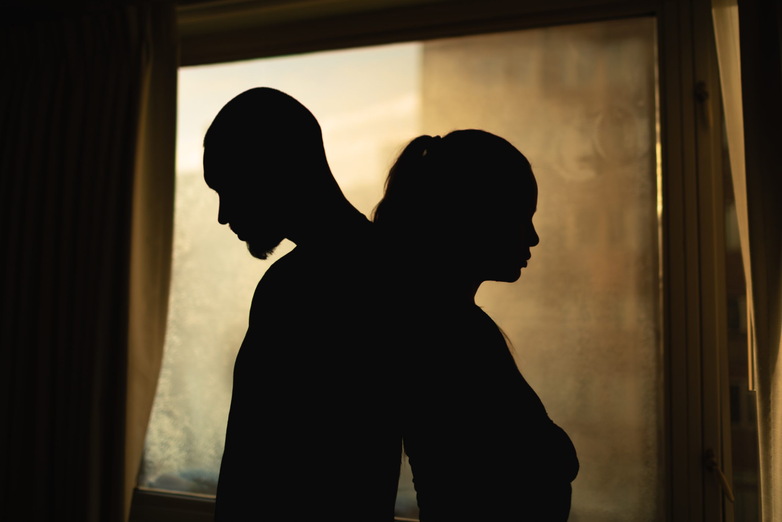 Silhouettes of two partners standing back-to-back in front of a window, facing away from one another, symbolizing emotional distance, unresolved conflict, and disconnection within a relationship.