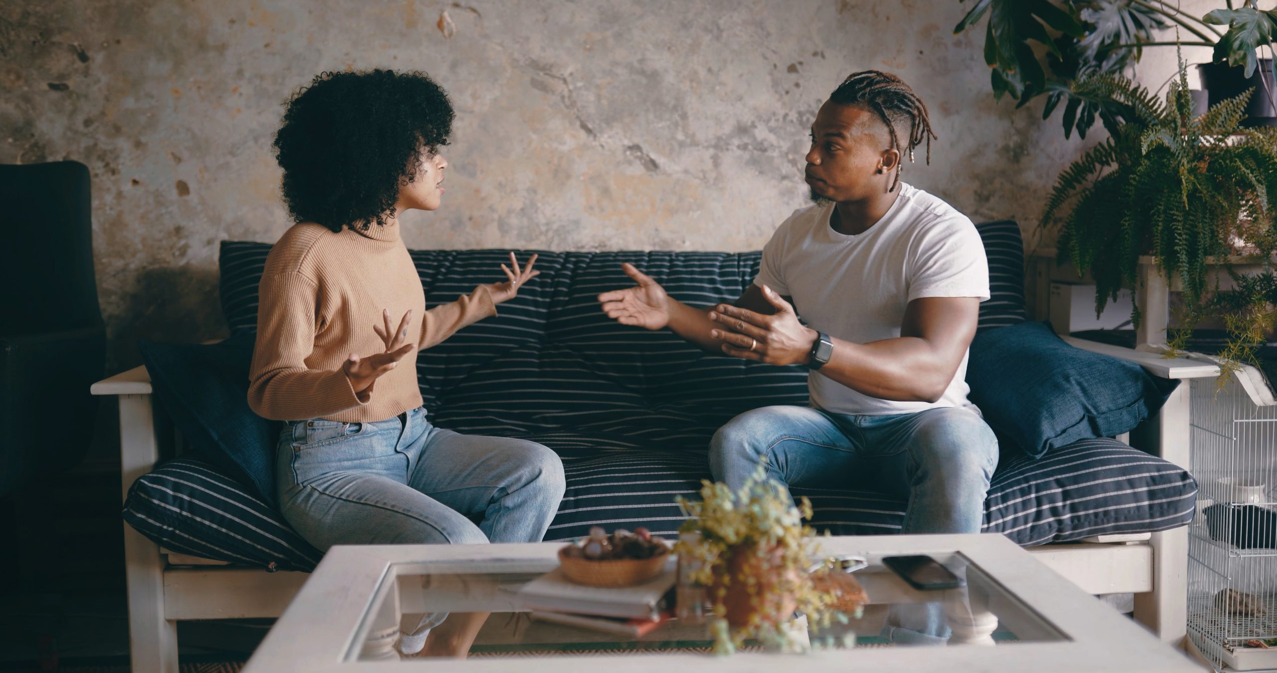 A couple sits on opposite ends of a couch engaged in a tense conversation, illustrating relationship conflict, communication challenges, and emotional disconnection.