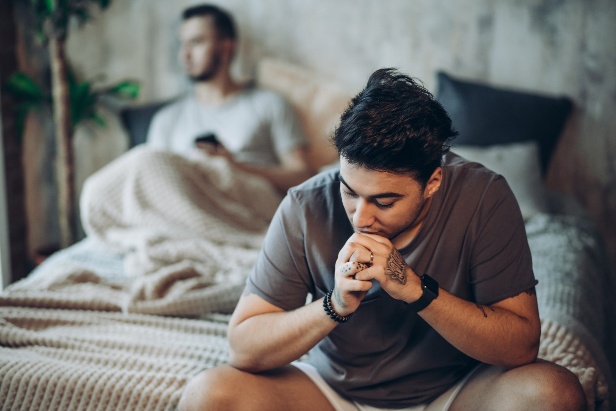 One partner sits in the foreground with their hands clasped in thought while another sits blurred in the background, illustrating emotional distance, uncertainty, and the aftermath of relationship betrayal.