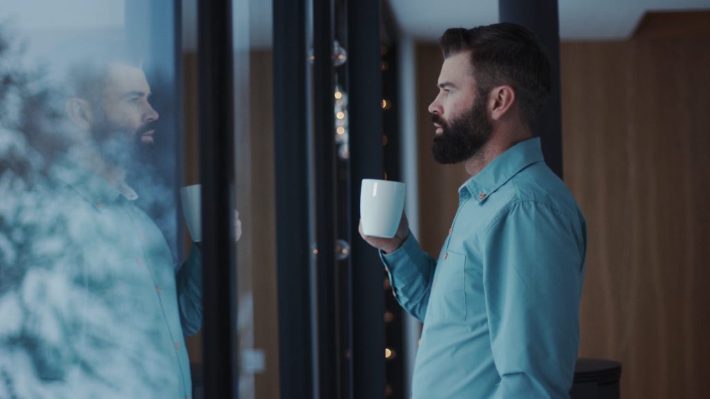 Person standing by a window holding a coffee mug and looking at their reflection, conveying introspection, self-reflection, and personal identity exploration.