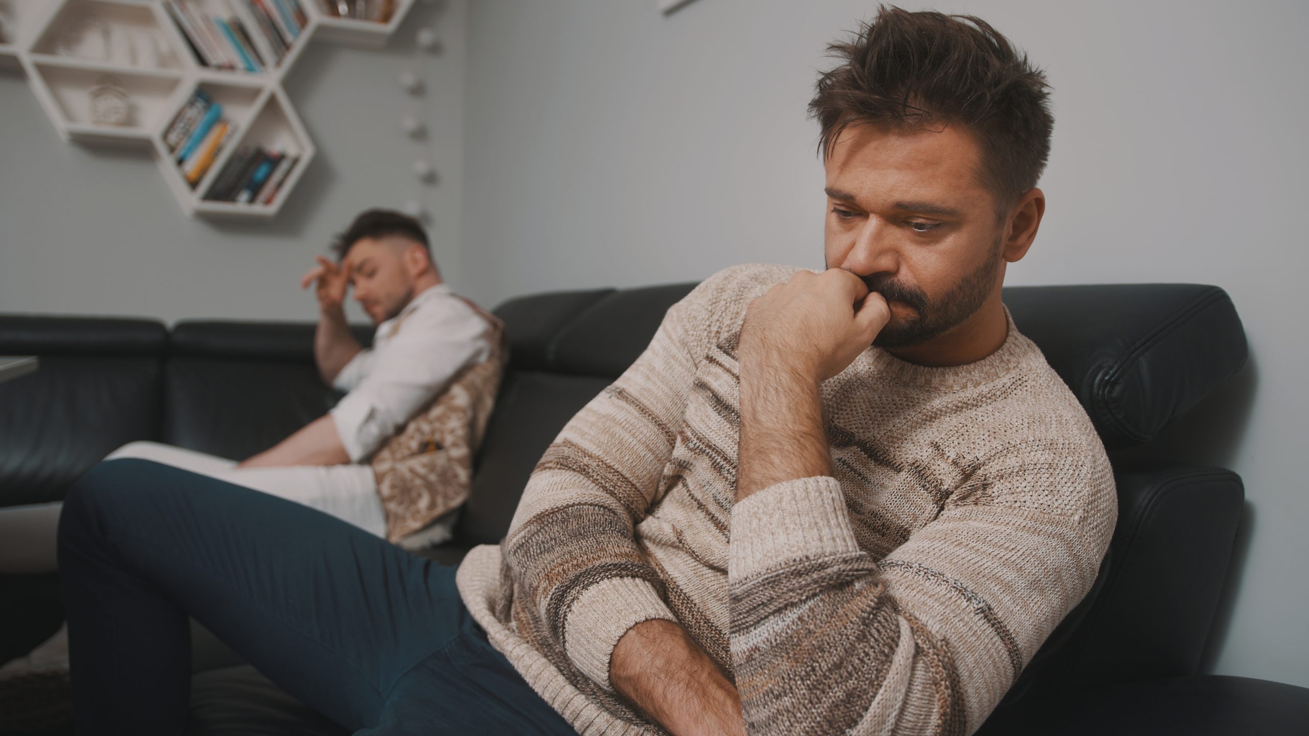 Two men sitting apart on a couch, both appearing distressed and emotionally disconnected after a conflict, illustrating relationship strain and unresolved tension.