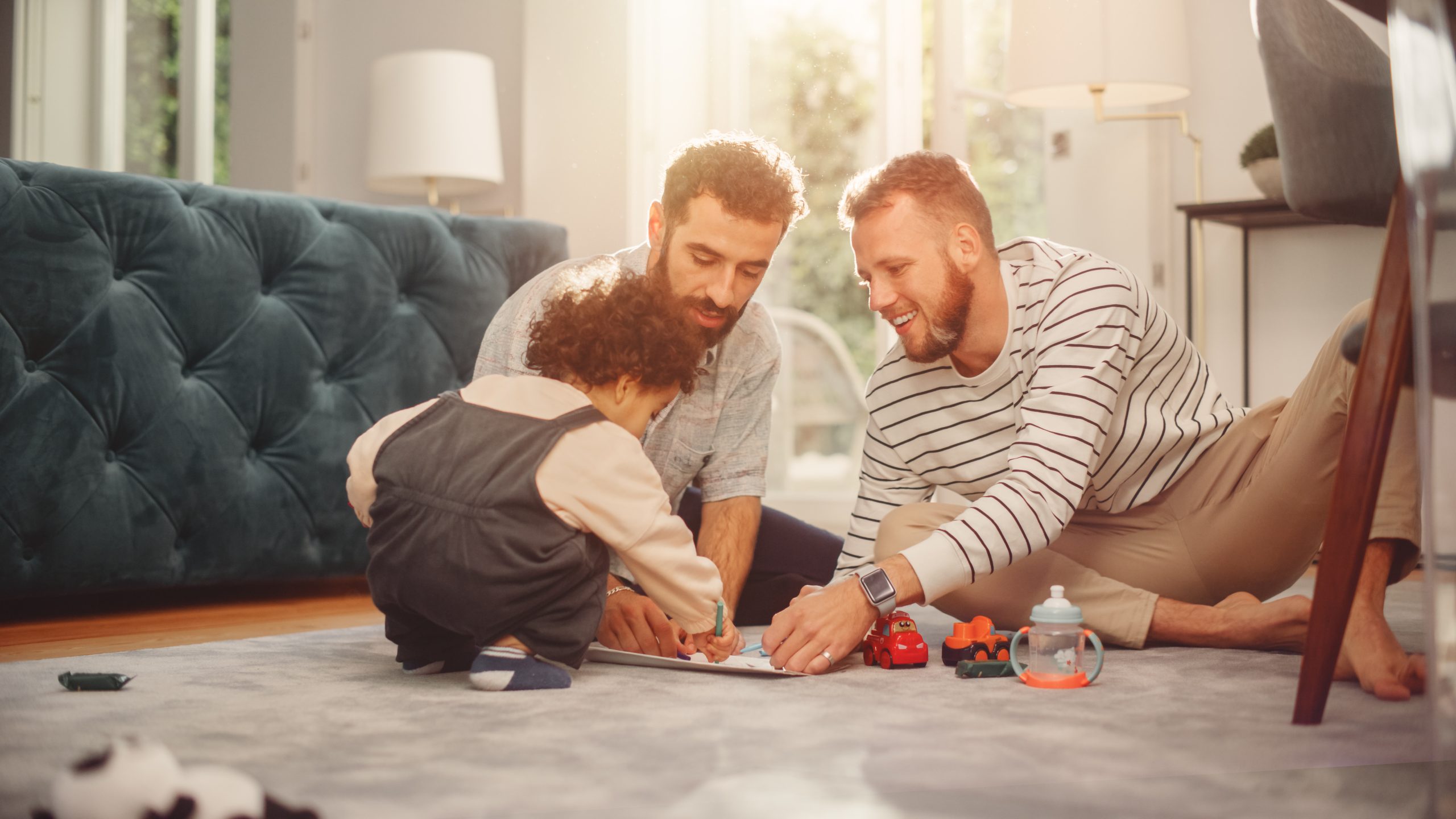 Same-gender couple and young child spending time together on the floor of their home, reflecting connection, family, and LGBTQ+ relationships.