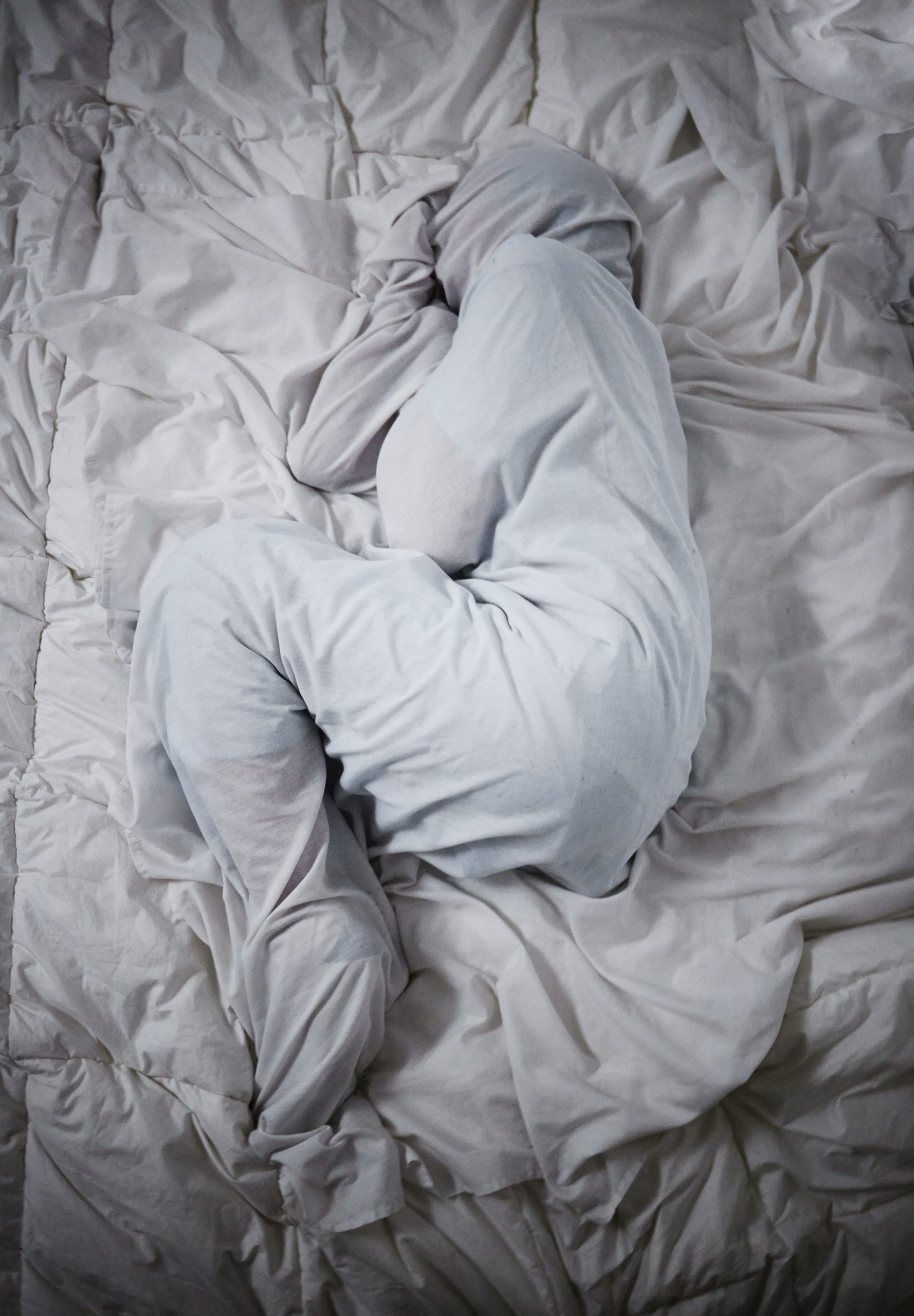 Person curled into a protective fetal position on a bed, wrapped in light-colored clothing and bedding, conveying emotional withdrawal, overwhelm, and the instinct to seek safety.