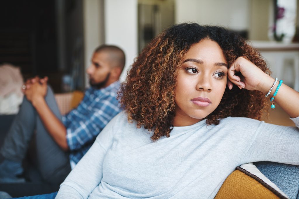 Person sitting in the foreground looking away with a distant, pained expression while their partner sits blurred in the background, illustrating emotional disconnection and relationship distress.